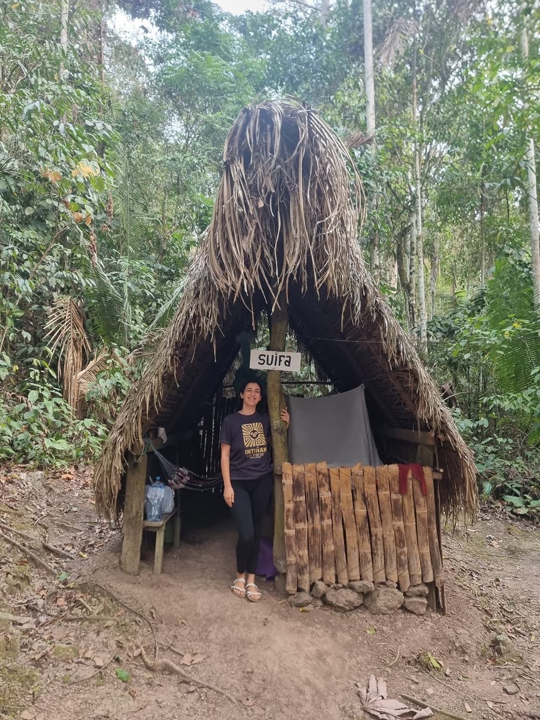 Asha Singh standing at the entrance of a thatched hut in the rainforest.